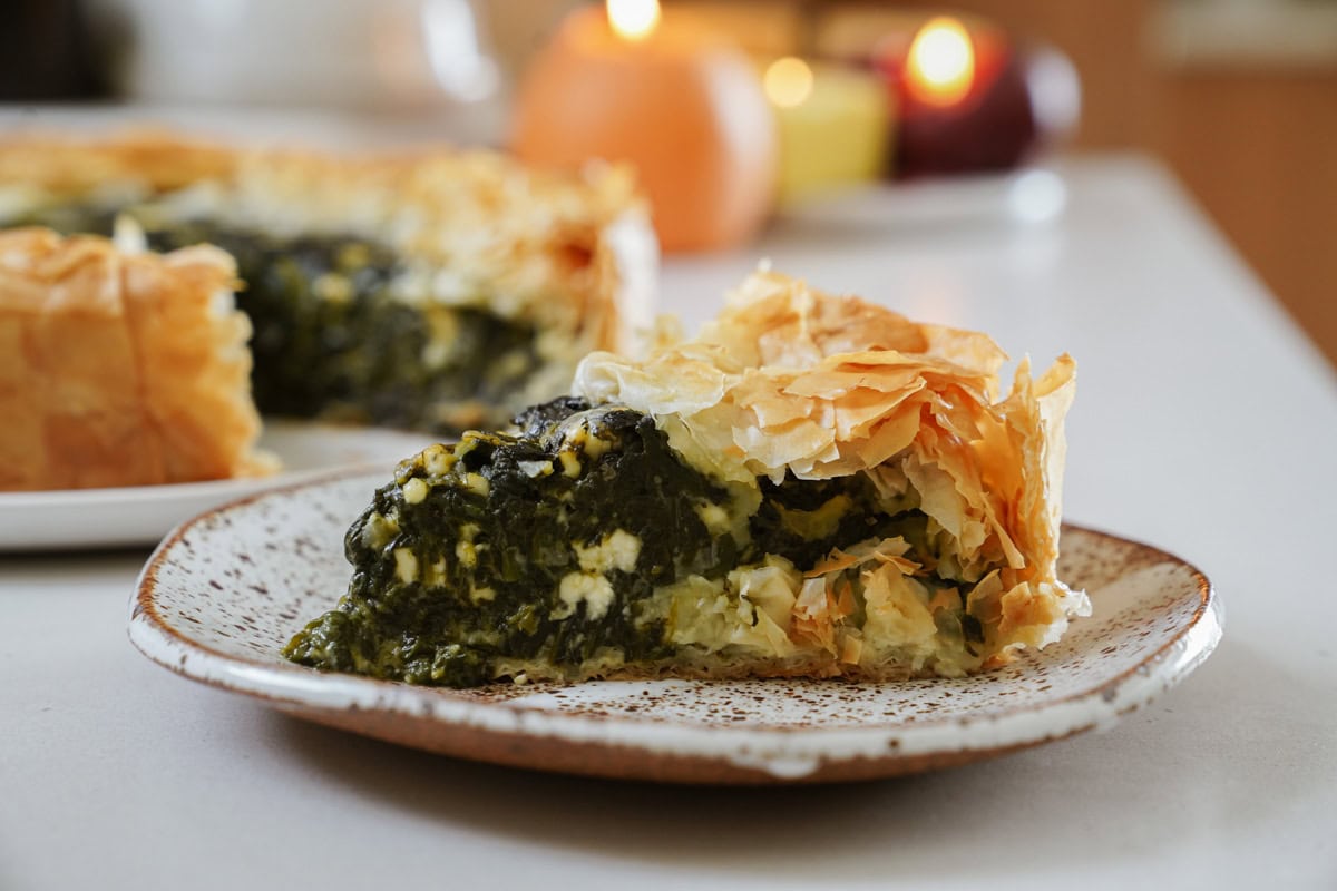 A slice of spinach and cheese pie with flaky, golden phyllo crust on a speckled plate. The pies rich green and creamy filling is visible. In the background, candles and a whole pie can be seen on a table.