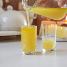 A hand pours freshly squeezed orange juice from a pitcher into one of two clear glasses on a kitchen counter. A white container and butter on a dish are blurred in the background.