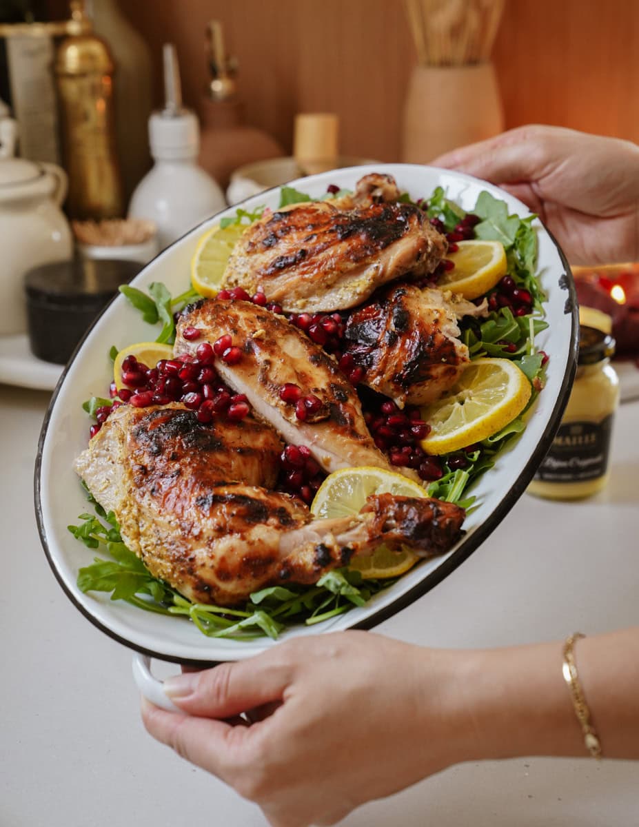 A person holding a white oval platter filled with spatchcock roasted chicken pieces on a bed of arugula, garnished with lemon slices and pomegranate seeds. The background shows kitchen items like a teapot and condiments.