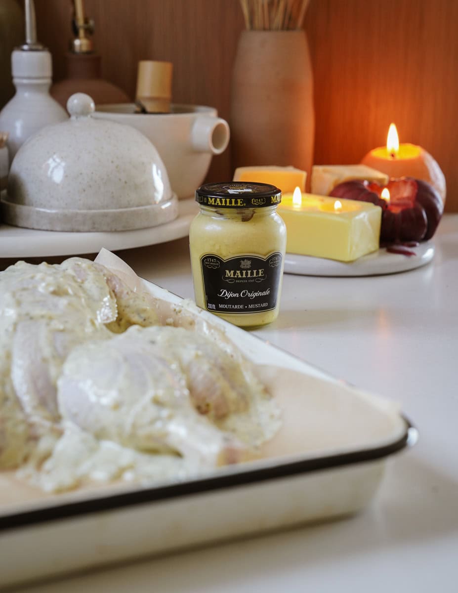 A kitchen scene showing raw chicken pieces coated in a creamy marinade on a baking tray. A jar of Dijon mustard, a teapot, lit candles, unpeeled red onions, and a block of butter are in the background.