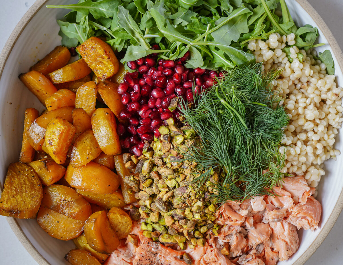 A colorful salad with arugula, golden roasted beets, pomegranate seeds, pearl couscous, dill, shredded salmon, and chopped pistachios, all arranged in sections within a large bowl.