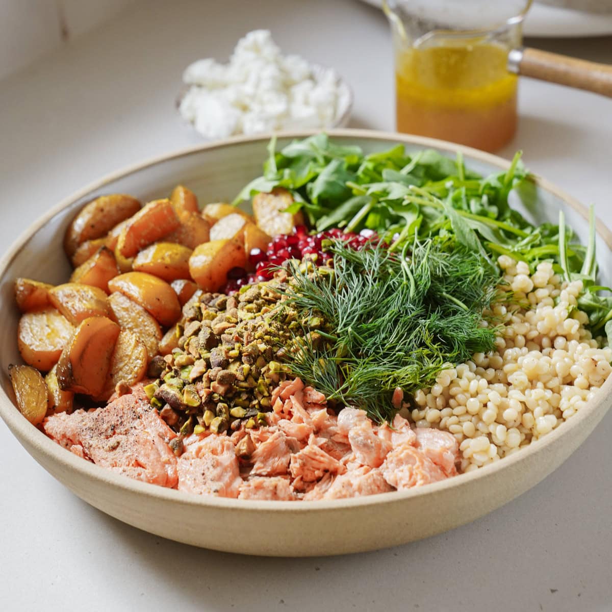 A bowl with salmon, roasted potatoes, arugula, couscous, dill, pistachios, and pomegranate seeds. A small bowl of cottage cheese and a jar of dressing are in the background on a countertop.