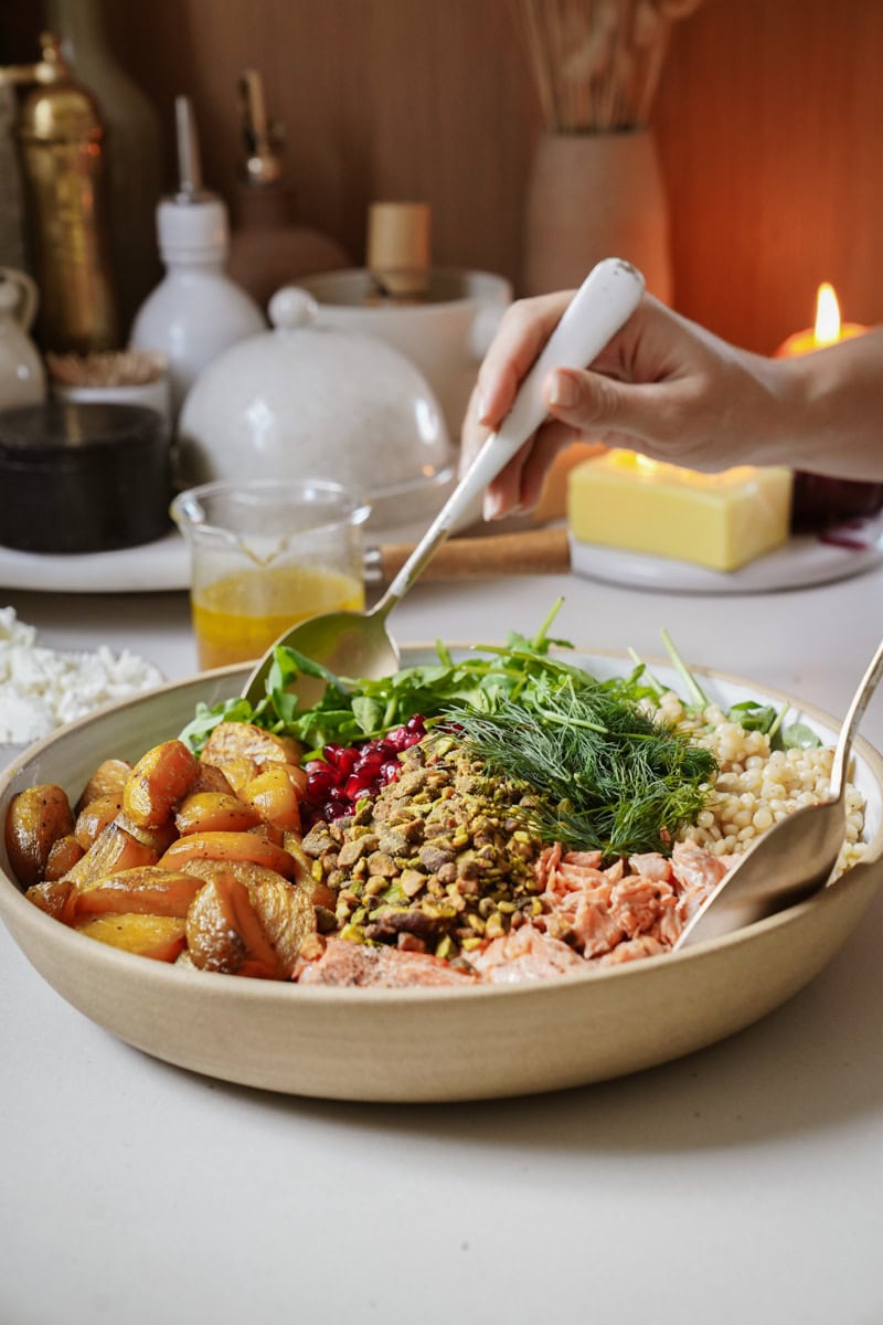 A hand with a fork reaches into a large bowl filled with a variety of colorful ingredients: roasted potatoes, pistachios, pomegranate seeds, greens, couscous, dill, and shredded salmon. In the background are condiments and a lit candle.