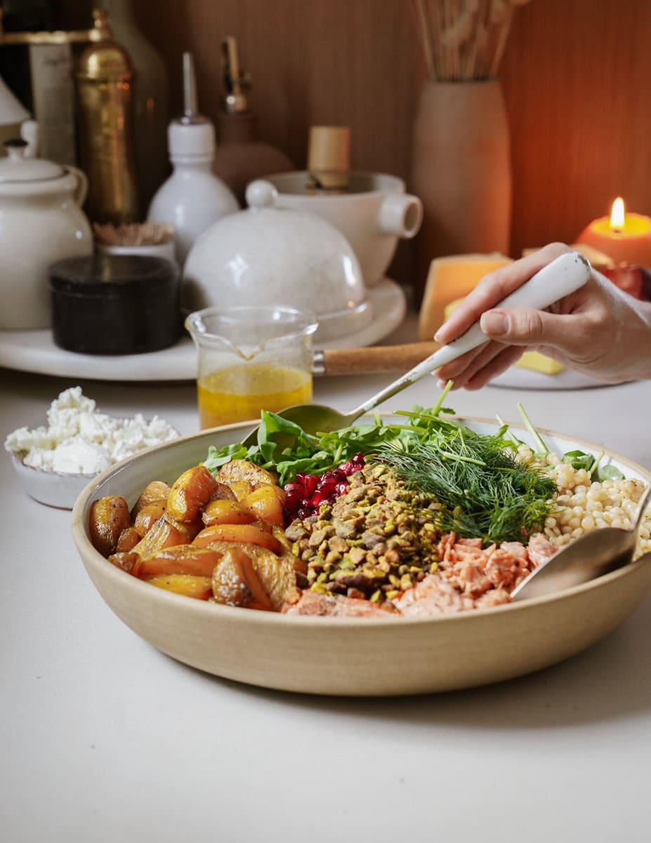 A bowl filled with ingredients like roasted potatoes, pomegranate seeds, pistachios, greens, and salmon. Theres a spoon in the bowl, a hand adding more ingredients, and kitchen items, including a lit candle, in the background.