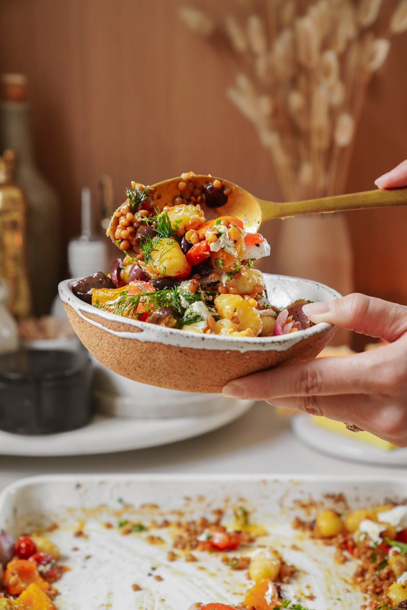 A person holds a ceramic bowl filled with a colorful salad, using a spoon to serve it. The salad includes grains, cherry tomatoes, olives, and fresh herbs, creating a vibrant mix. A kitchen setting is visible in the background.