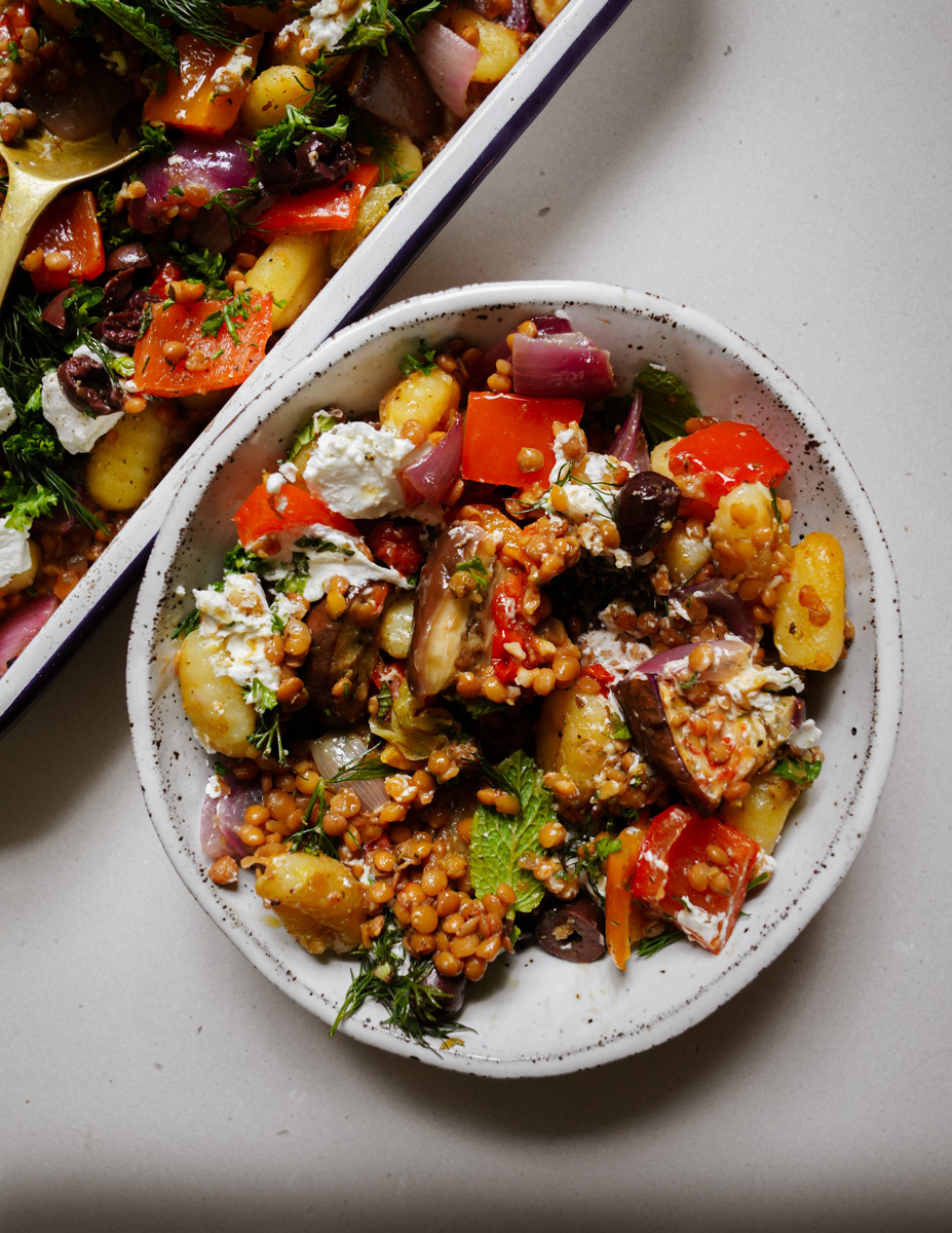 A plate of colorful Mediterranean salad with ingredients like gnocchi, red peppers, onions, olives, lentils, and feta cheese. A larger dish with the same salad is partially visible above the plate.