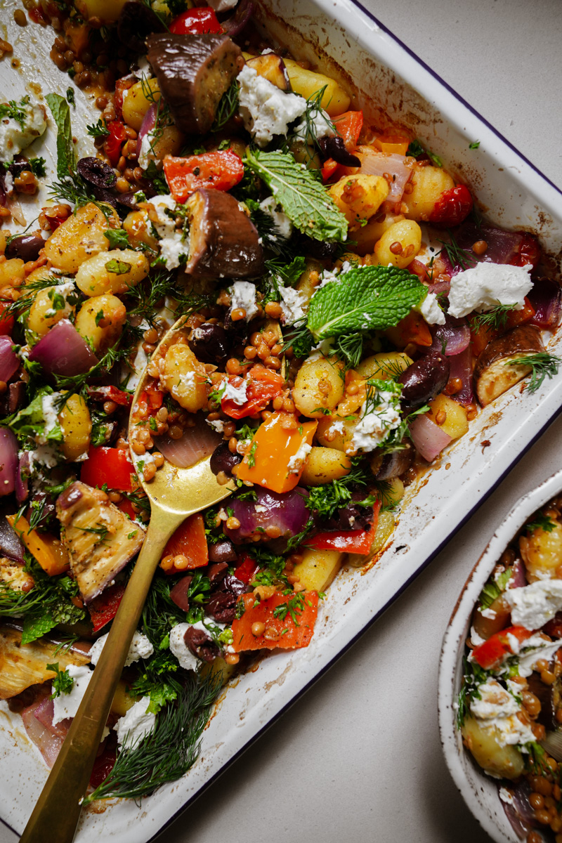 A colorful roasted vegetable and gnocchi dish in a baking tray. It includes bell peppers, red onions, eggplant, and leafy greens, topped with dollops of white cheese. A brass serving spoon is placed on the tray.