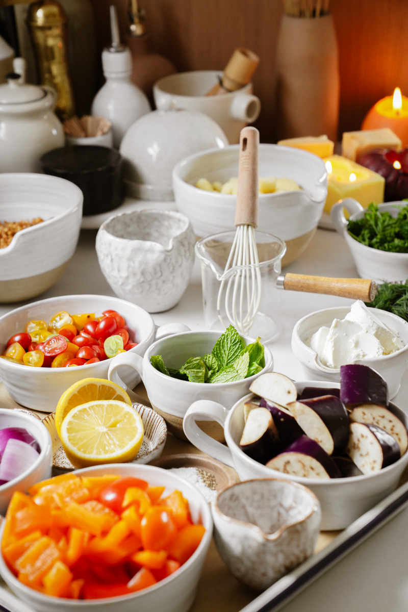 A variety of fresh ingredients arranged on a table, including sliced cherry tomatoes, lemon halves, eggplant, bell peppers, mint leaves, and bowls of cream and herbs. A whisk is placed in the center alongside bowls and a candle in the background.