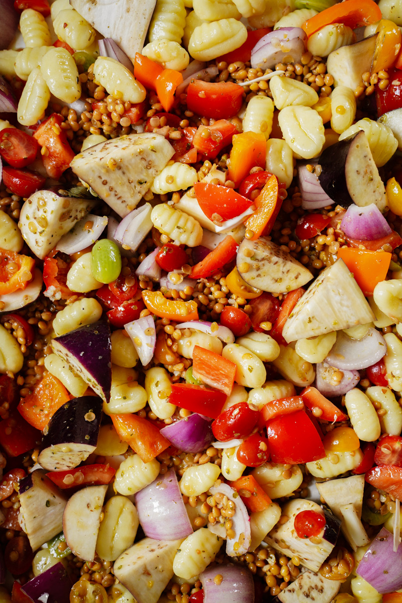 A colorful salad with ingredients like gnocchi, cherry tomatoes, eggplant, bell peppers, red onions, corn, and grains mixed together, showcasing a variety of textures and vibrant colors.