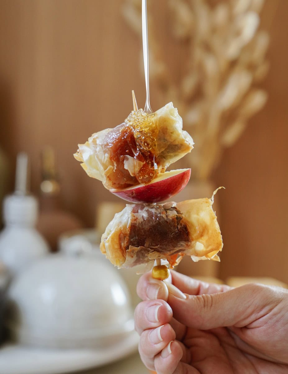 A hand holds a skewer with baked goods, apple slices, and dripping honey against a blurred background with warm tones.