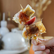 Close-up of a hand holding a skewer with two pieces stuffed dates. The background is softly blurred, creating a warm, inviting atmosphere.