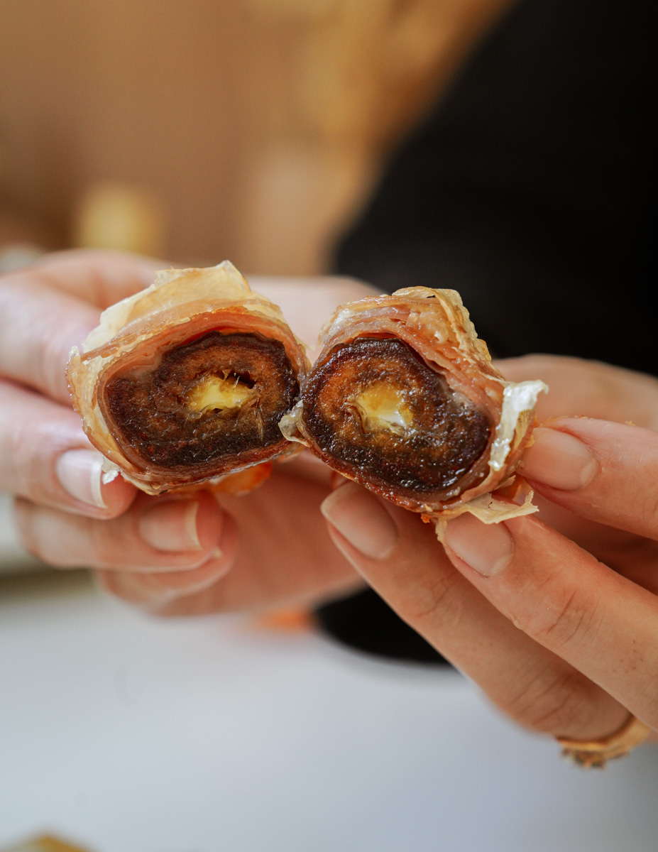 Close-up of hands holding a halved date wrapped in bacon, revealing a creamy filling inside the dark brown fruit. The hands have light-colored nail polish, and the background is softly blurred.