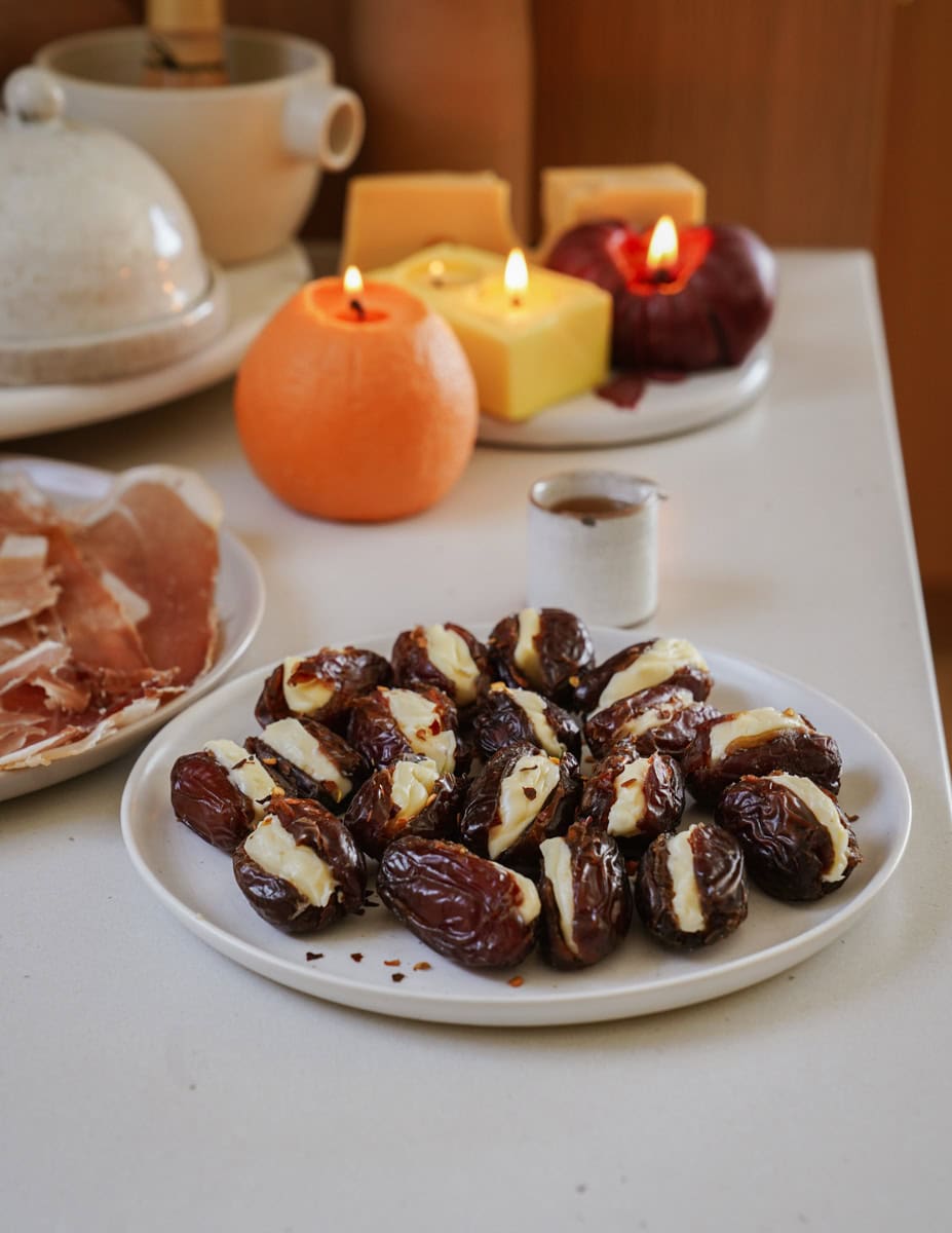 A plate of dates filled with cheese and sprinkled with red pepper flakes sits on a white surface. Nearby are lit candles, a peeled orange, slices of cured meat, and a small cup, creating a warm and cozy setting.