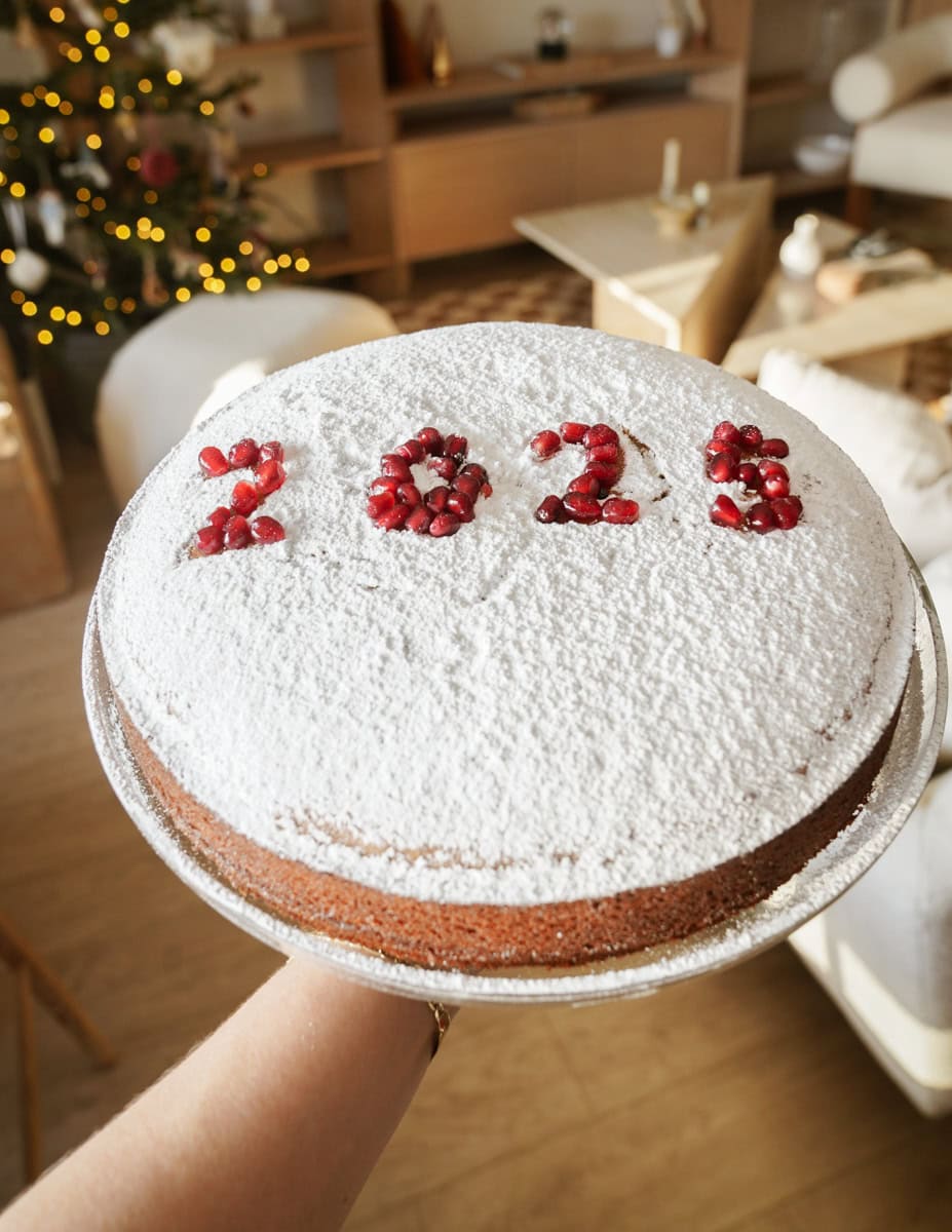 A hand holds a cake dusted with powdered sugar, featuring the numbers 2023 written with pomegranate seeds. A Christmas tree with lights is visible in the cozy living room background.