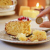 A slice of Vasilopita with pomegranate seeds on top sits on a speckled plate. A hand is holding a one euro coin near the cake. A fork is on the plate with crumbs scattered around. Candles and a blurred background create a cozy atmosphere.