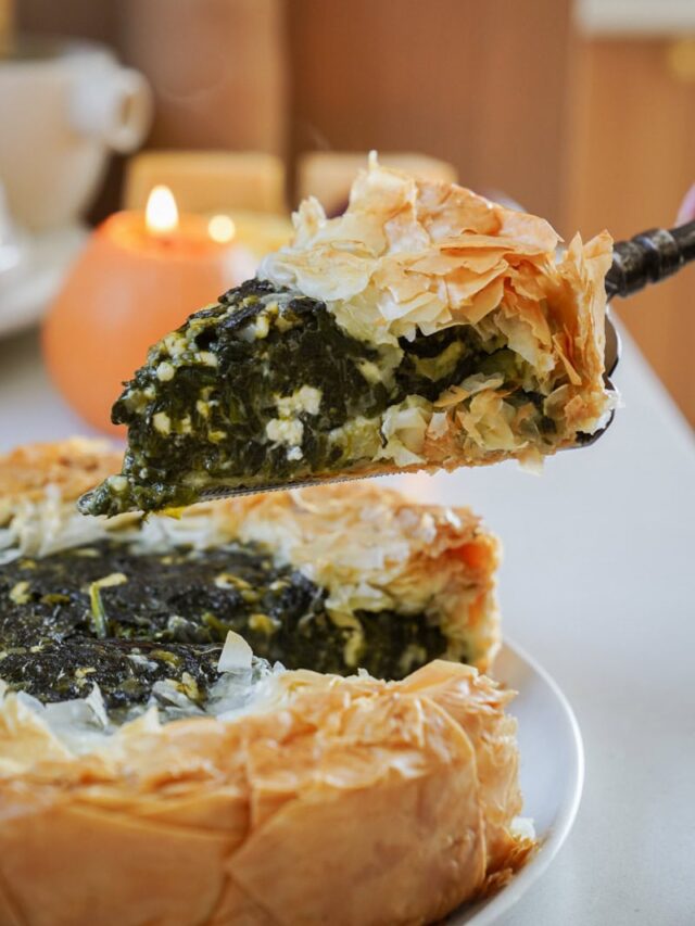 A slice of spinach pie with flaky crust is being lifted from a whole pie on a plate. The pie has a rich green spinach filling with hints of cheese and a golden-brown crust. In the background, there are soft-focus candles.