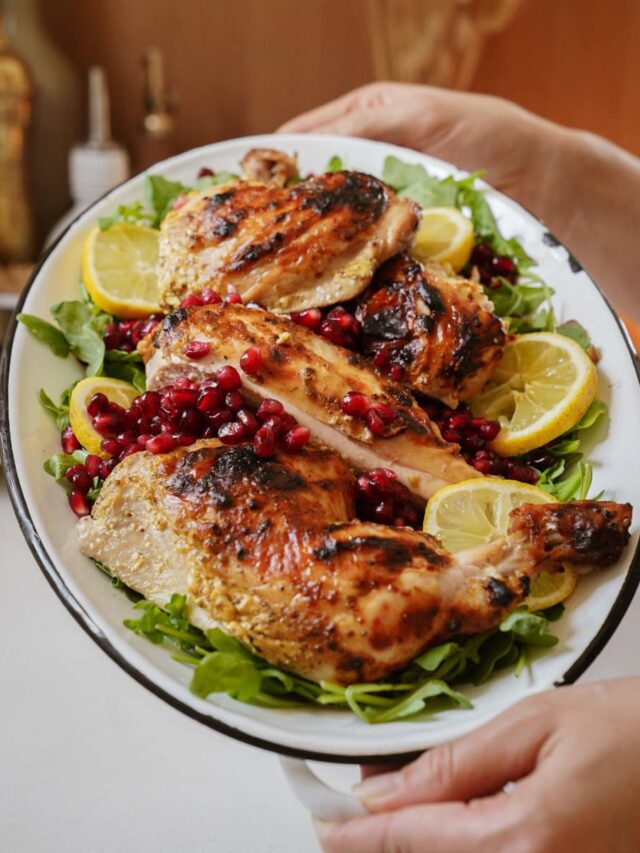 Plate of grilled chicken pieces on a bed of greens, garnished with pomegranate seeds and lemon slices, held in hands.