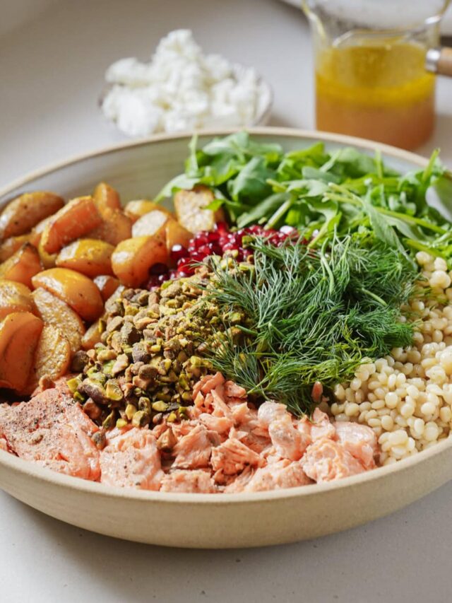 A bowl filled with roasted potatoes, arugula, couscous, fresh dill, flaked salmon, pistachios, and pomegranate seeds. A small bowl of soft cheese and a glass container of dressing are in the background.