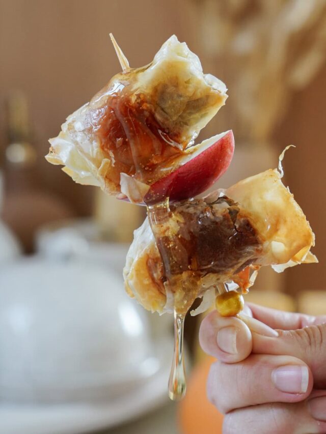 Close-up of a hand holding a skewer with two pieces of crispy, glazed pastry wrapped around fruit, likely figs or similar, with a light drizzle of honey or syrup. The background is softly blurred, focusing on the skewered treat.