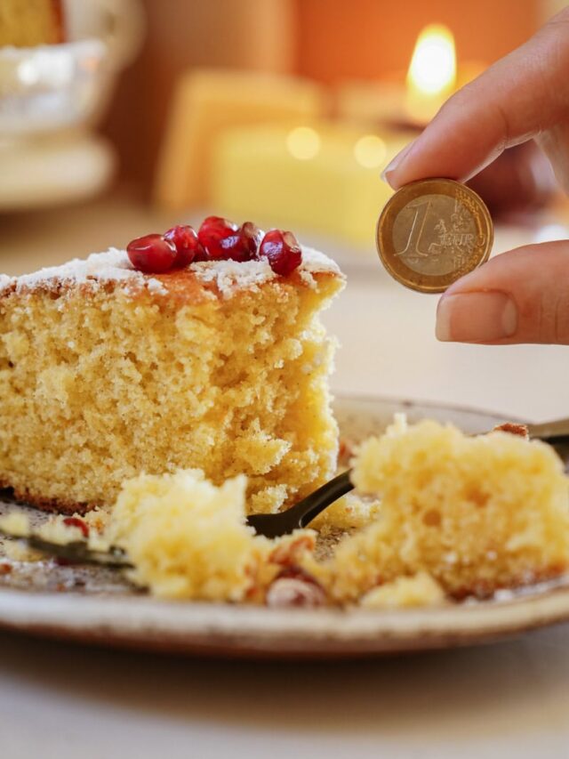 A slice of yellow cake topped with pomegranate seeds on a plate. A hand holding a one euro coin is positioned next to the cake. There is a lit candle in the blurred background.