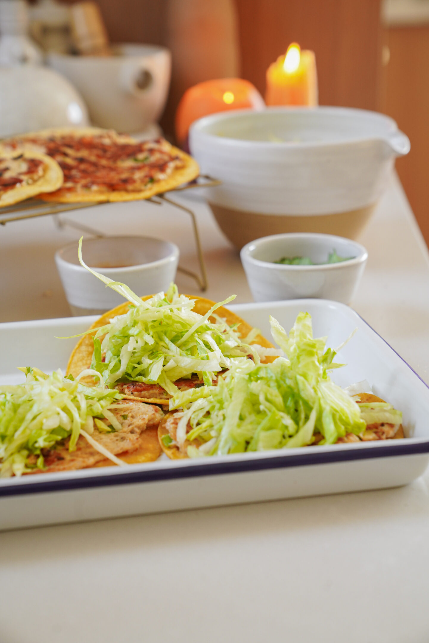 Tacos topped with shredded lettuce in a white dish are on a counter alongside small bowls and a lit candle. Another dish with what appears to be pizza is in the background.