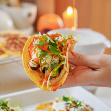 A hand holds a taco filled with vibrant ingredients like lettuce, cilantro, and diced vegetables. A bright orange sauce is being drizzled over the top. A blurred background features a glowing candle and more food items.