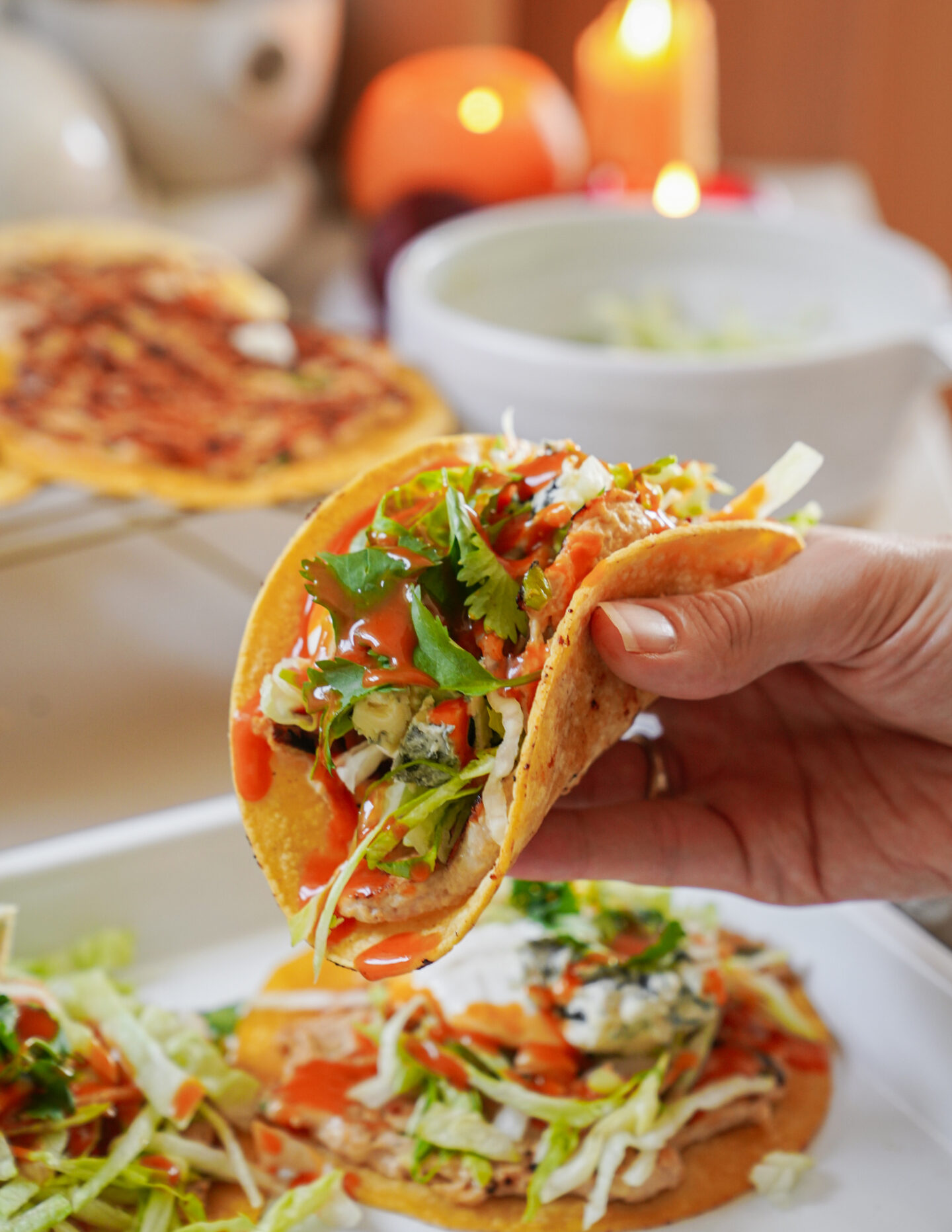 A person holding a taco filled with shredded lettuce, cilantro, and white crumbled cheese, topped with red sauce. More tacos are on a tray in the background, and theres a lit candle nearby.