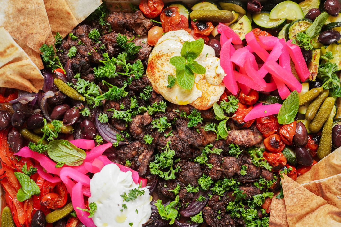 A colorful platter featuring spiced grilled meat topped with parsley, surrounded by pickled vegetables, pink pickled onions, olives, roasted tomatoes, pita bread, dollops of hummus and yogurt, garnished with fresh herbs.