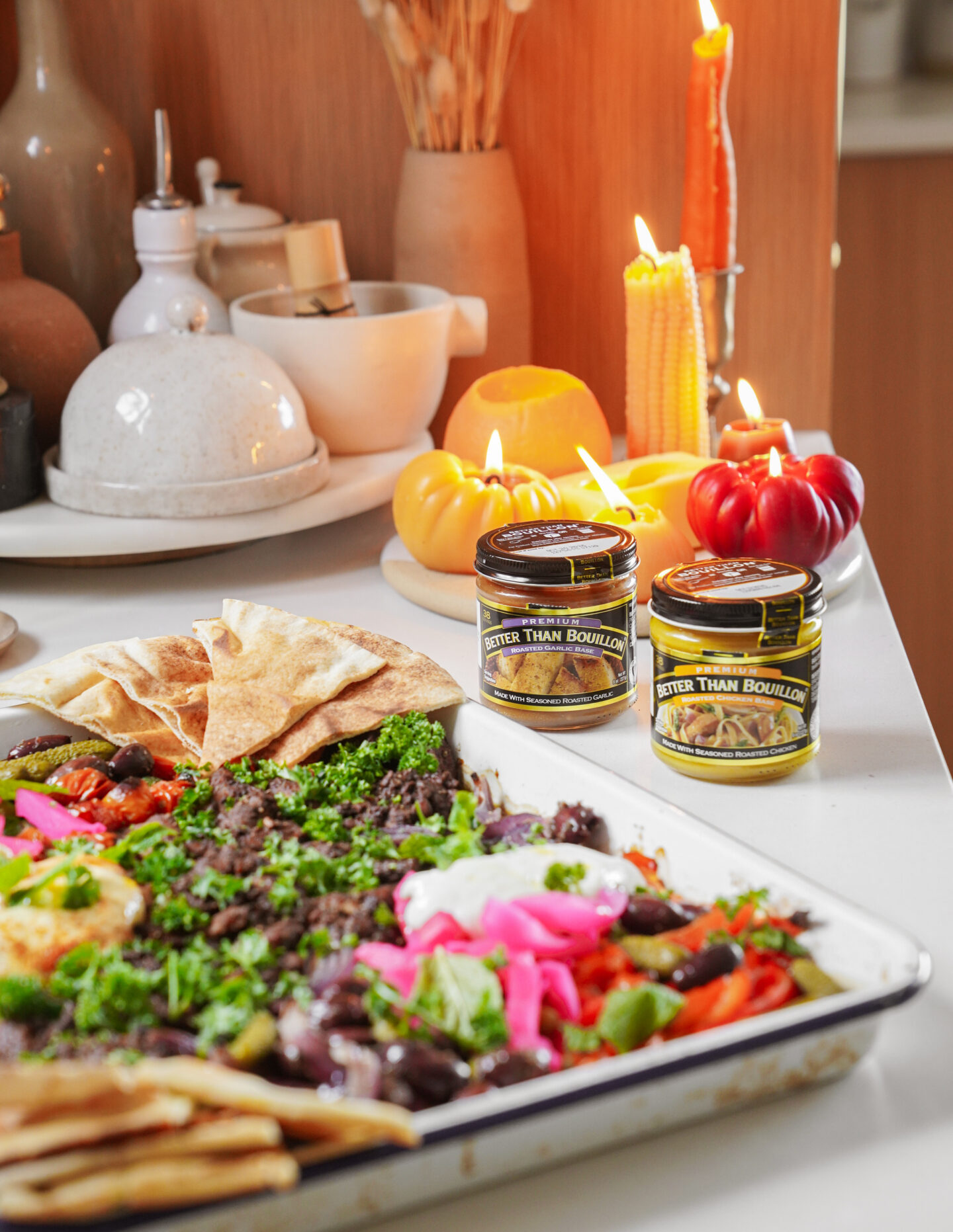 A platter of Mediterranean dips with pita bread, garnished with herbs and colorful vegetables. Two jars labeled Better Than Bouillon sit nearby. Candles and decorative pumpkins add a cozy touch in the background.