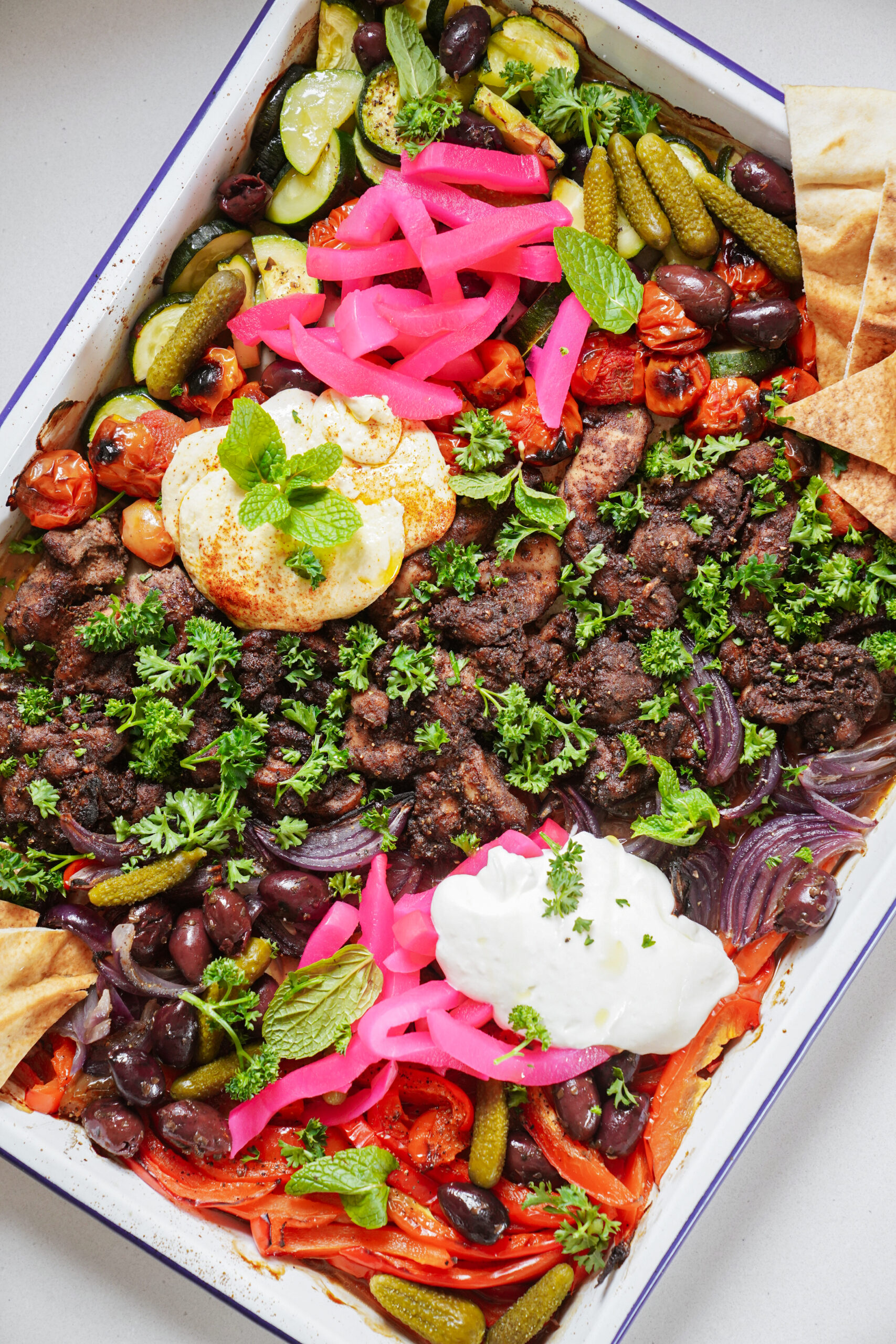 A colorful platter featuring grilled meat, vibrant pickled pink onions, olives, zucchini, cucumbers, roasted tomatoes, herbs, yogurt, and pita bread. Its arranged in a large dish, garnished with sprigs of mint and parsley.