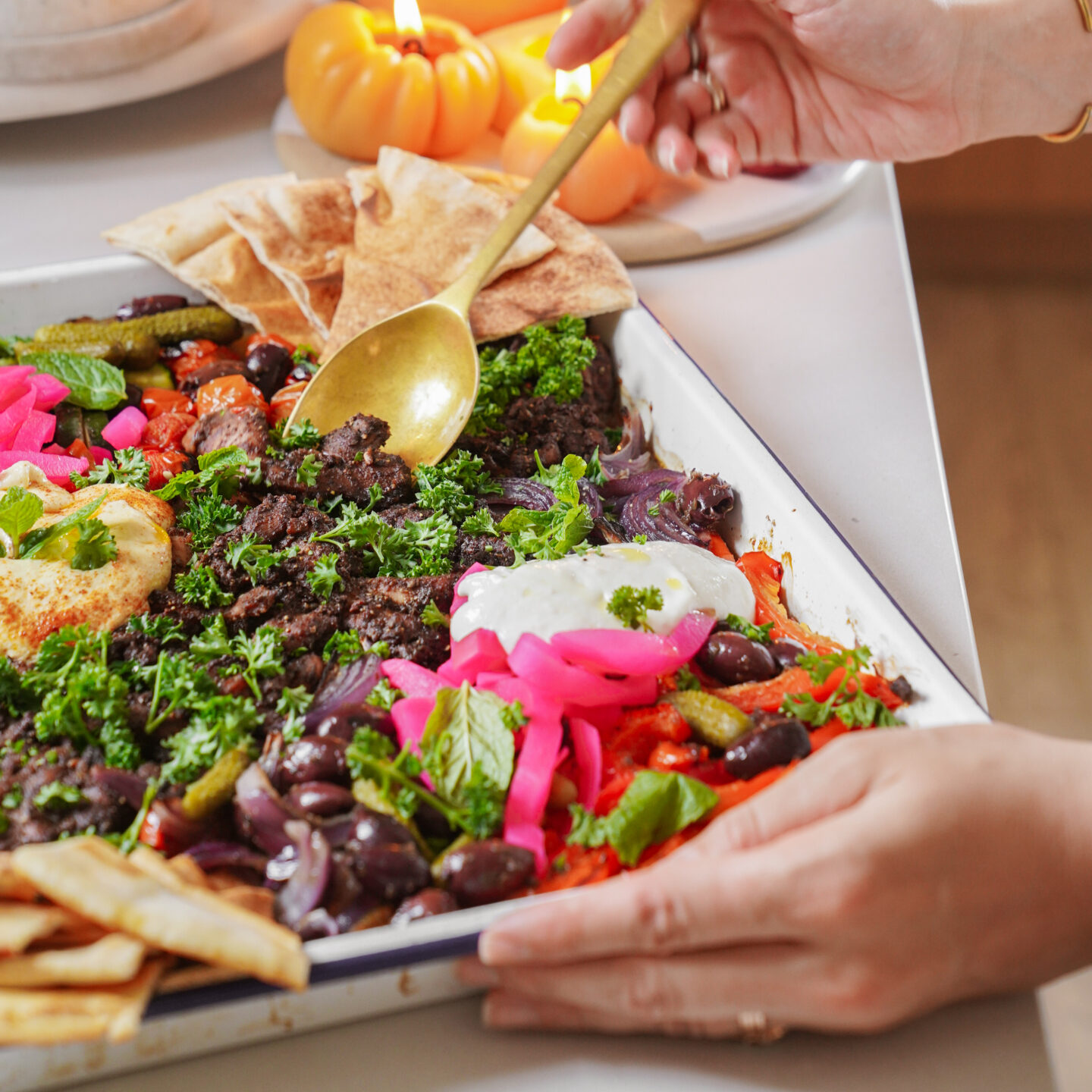 A person serves a roasted vegetable and black bean traybake with vibrant toppings like pink pickled onions, fresh parsley, yogurt, and pita bread. Small decorative pumpkins and lit candles are in the background.