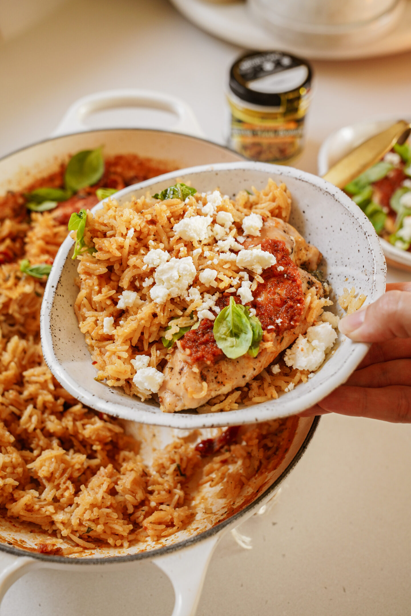 A person holds a bowl of chicken and rice garnished with herbs and crumbled cheese. The bowl is above a large pot filled with the same dish. Theres a jar of seasoning in the background on a kitchen counter.