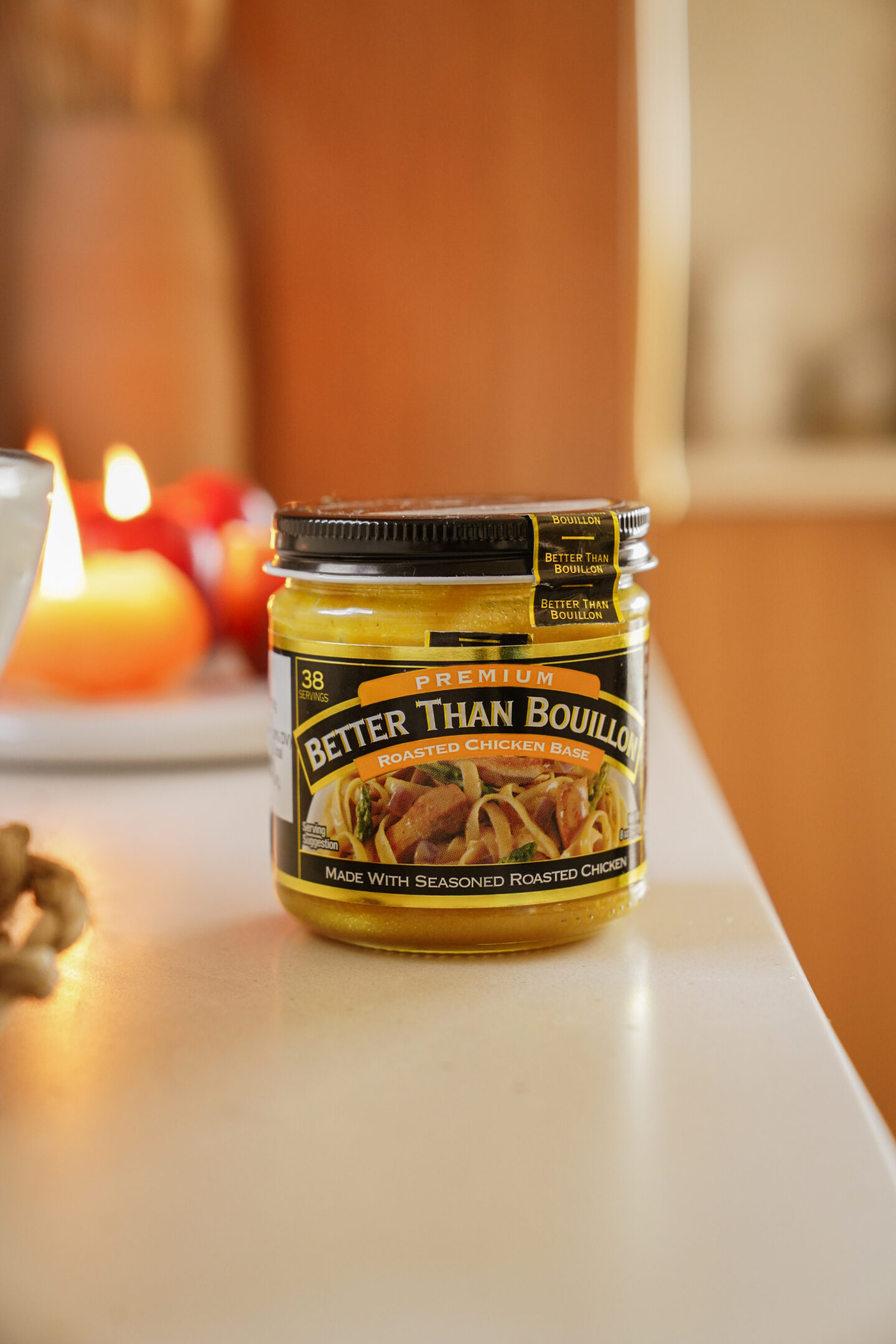 A jar of Better Than Bouillon roasted chicken base sits on a kitchen counter. The label is black and yellow with images of roasted chicken. In the background, there are blurred kitchen items and a glowing candle.