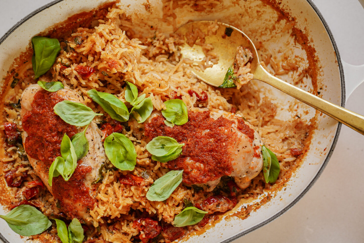 A baked dish featuring chicken breasts topped with tomato sauce, placed on a bed of roasted rice and sun-dried tomatoes. Fresh basil leaves are scattered on top. A gold spoon rests inside the dish.