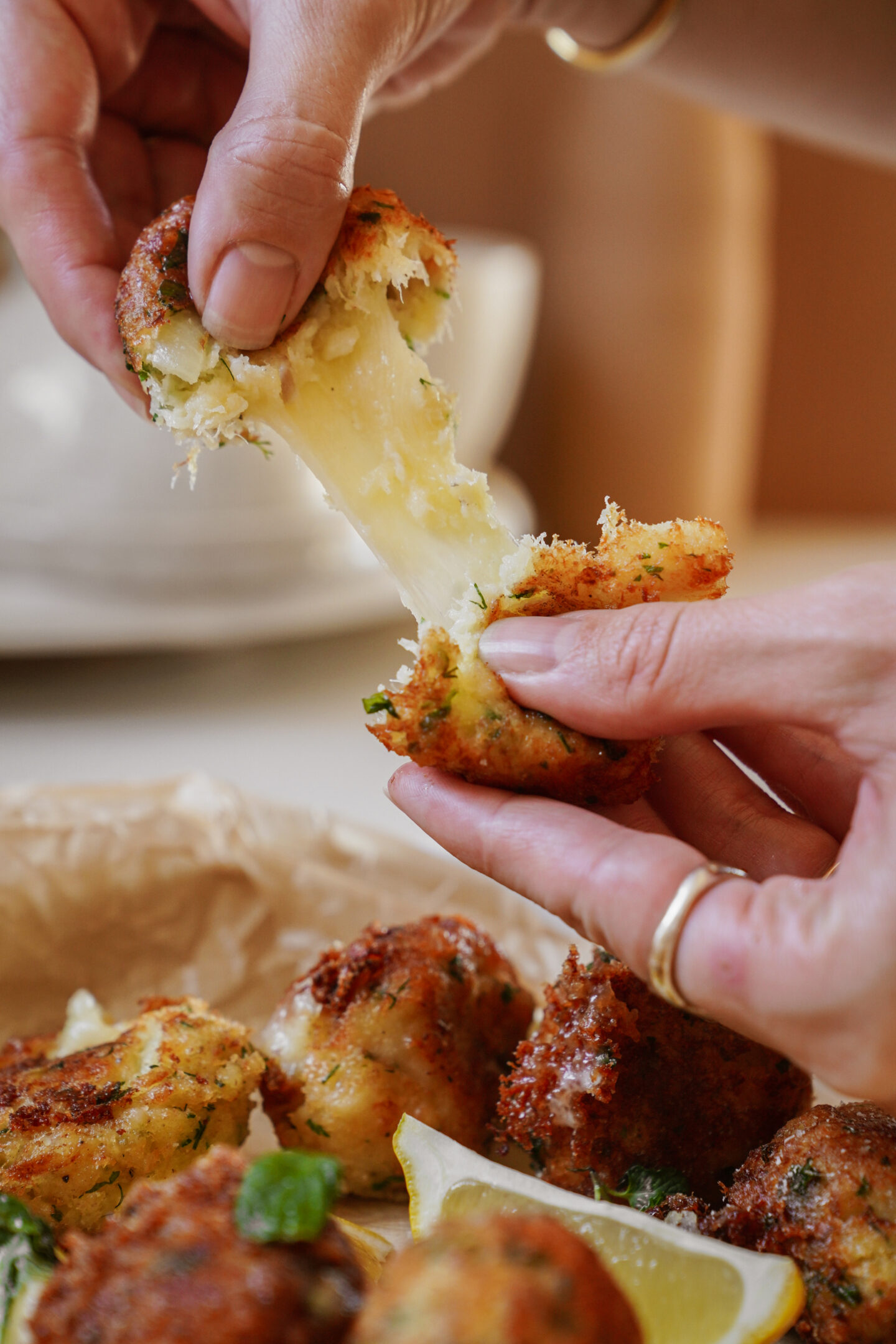 Close-up of hands pulling apart a crispy, fried cheese-filled ball, revealing melted cheese. The snack is garnished with herbs, and lemon wedges are visible on the side, with more snacks in the background on a tray.