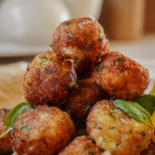 A close-up of a plate stacked with golden-brown, crispy croquettes garnished with fresh green herbs. The croquettes appear to have a crunchy texture, with visible flecks of herbs and breadcrumbs.