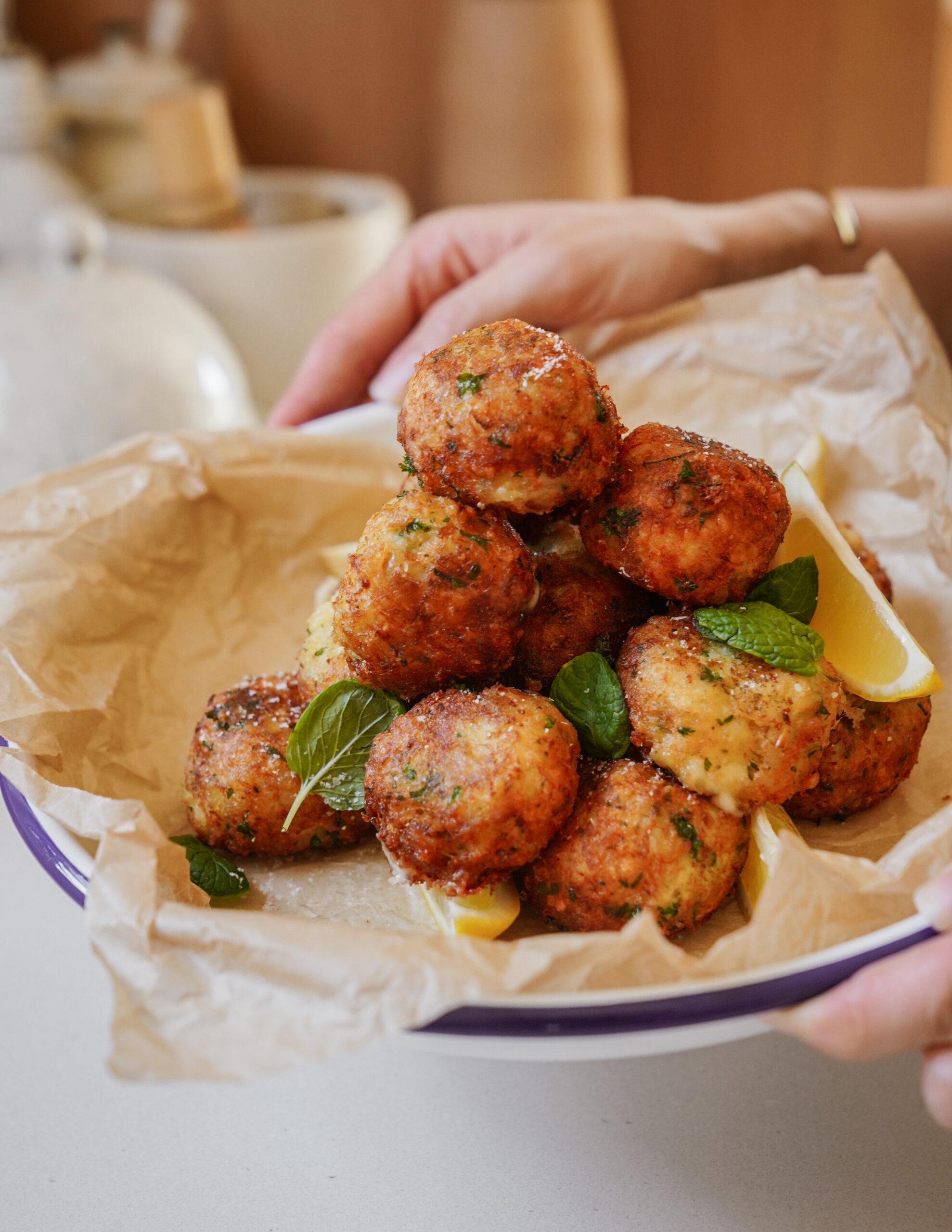 A plate of golden-brown, crispy arancini balls piled on parchment paper, garnished with fresh mint leaves and lemon wedges. The hands are holding the plate on a light-colored countertop.