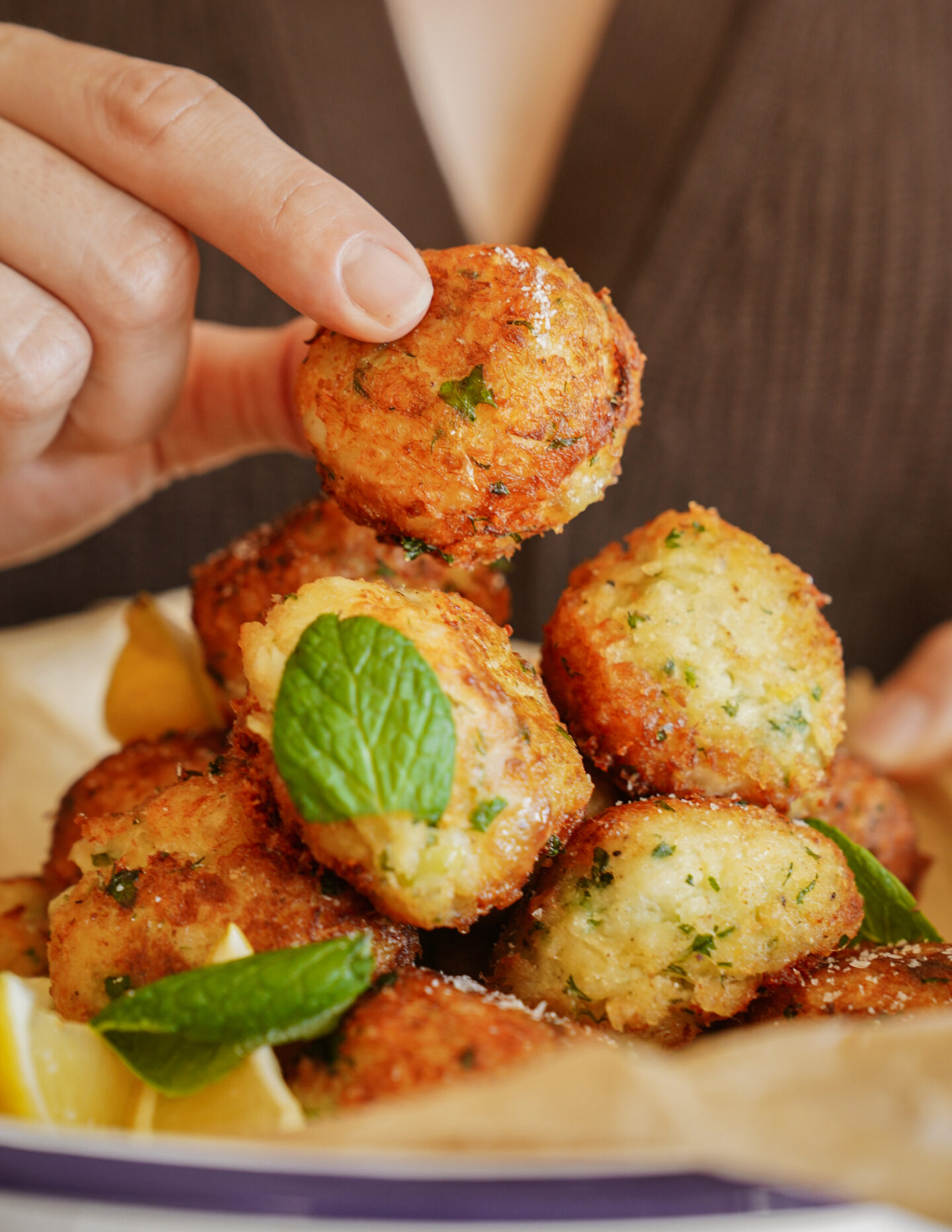 A hand picking up a golden, crispy fritter from a plate. The fritters are garnished with fresh herbs and accompanied by lemon wedges, creating an appetizing and fresh presentation.