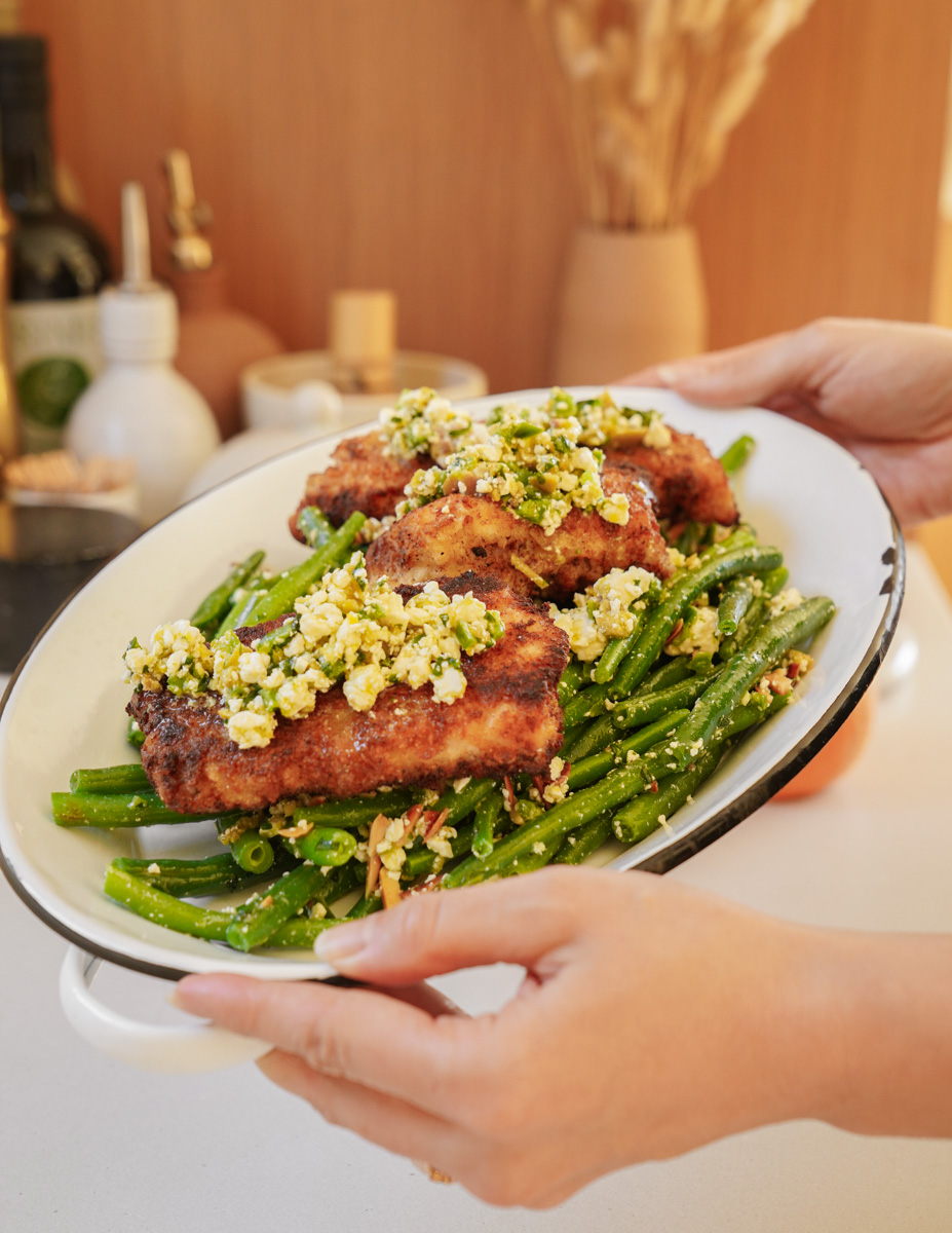 Hands holding a platter with crispy fried chicken topped with herbs served over a bed of sautéed green beans. In the background, a blurred kitchen setting with bottles and a vase.