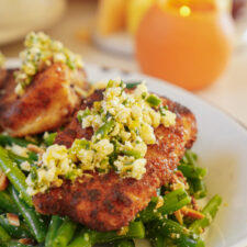 A close-up of a dish featuring a piece of seasoned fish topped with a chunky feta and herb mixture. Its served on a bed of vibrant green beans, garnished with sliced almonds. The background shows an orange candle adding warmth to the scene.