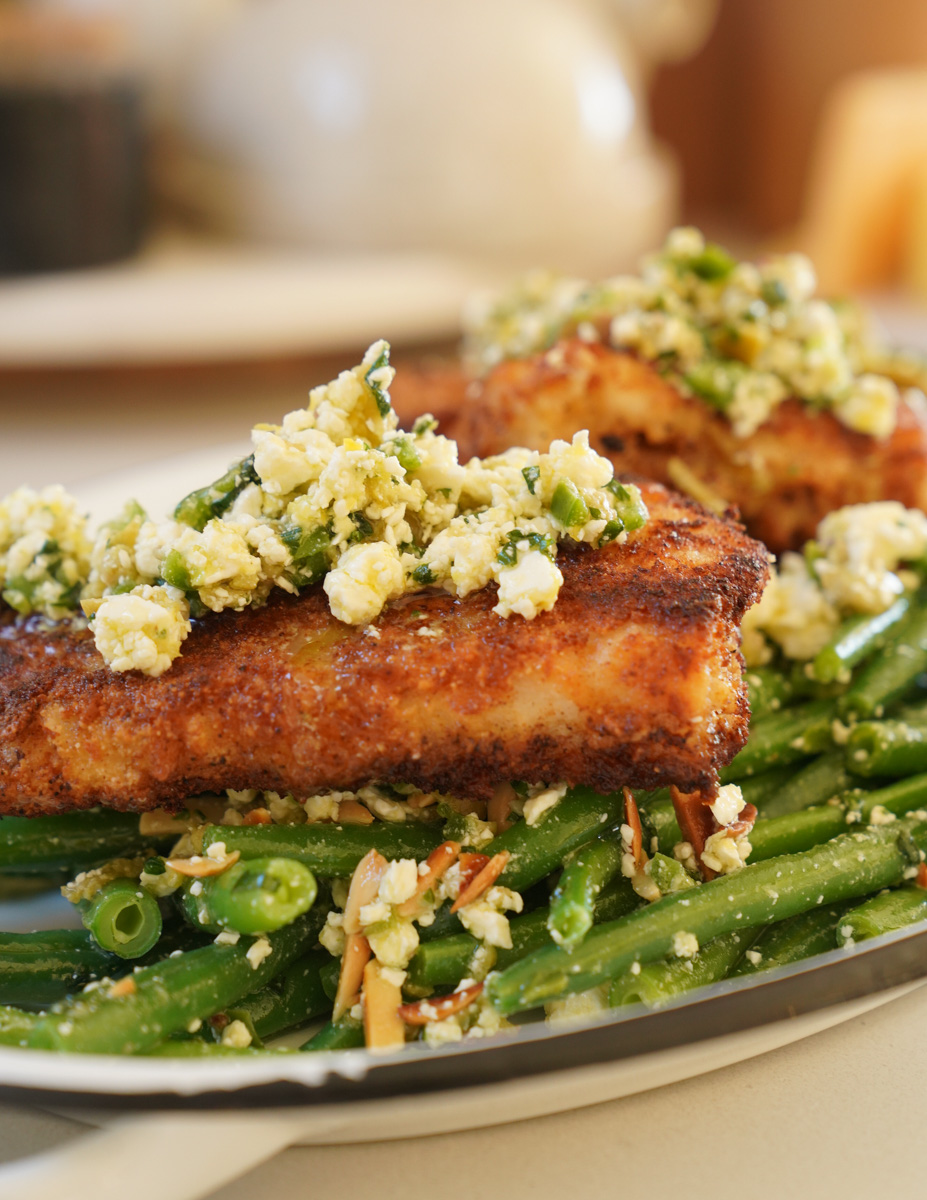 A plate of crispy salmon topped with herbed feta cheese, served on a bed of green beans garnished with chopped almonds and herbs. The dish is presented on a metal plate.