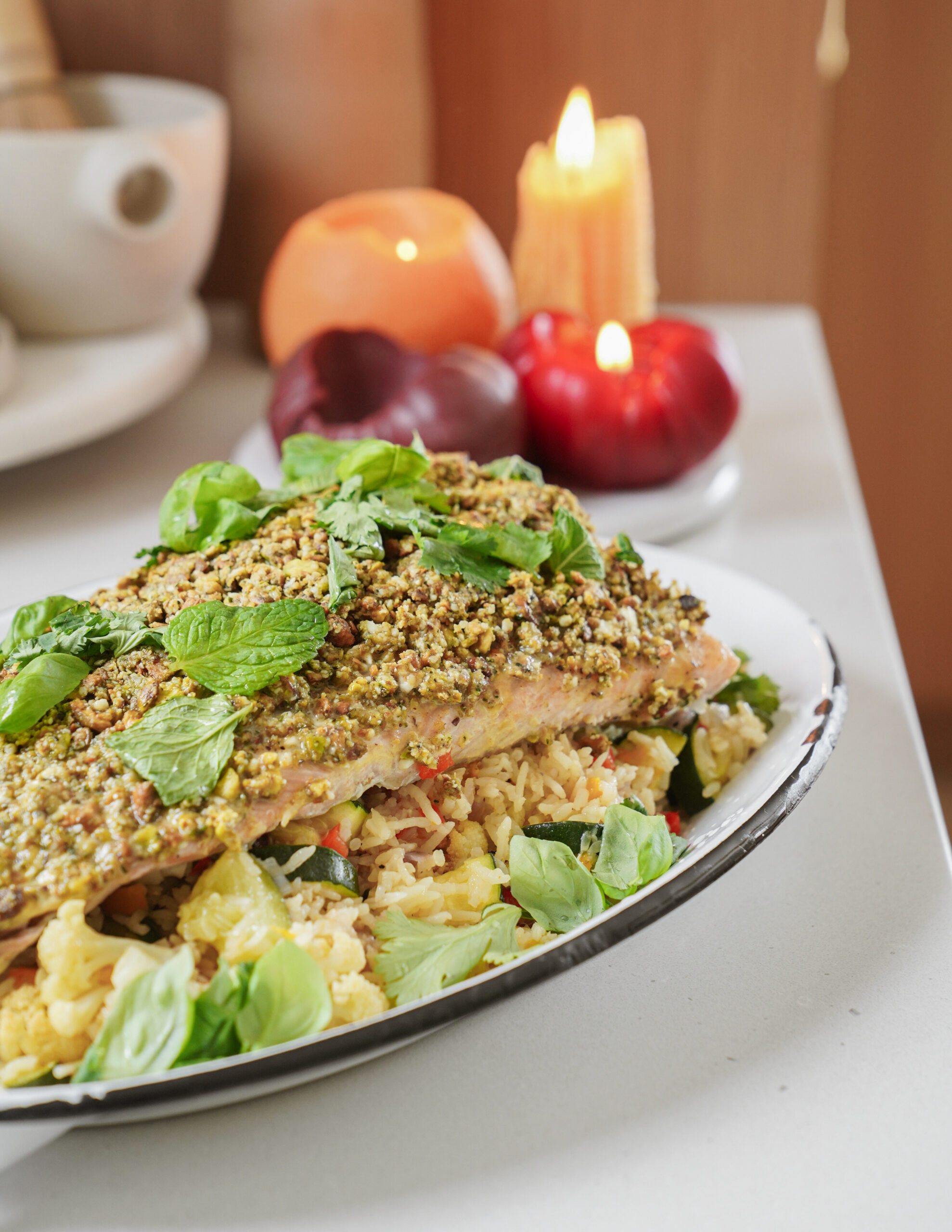 A plated dish featuring salmon topped with herbs and seeds, served on a bed of rice and vegetables. Fresh basil leaves garnish the meal. In the background, candles and decorative items add a cozy ambiance.