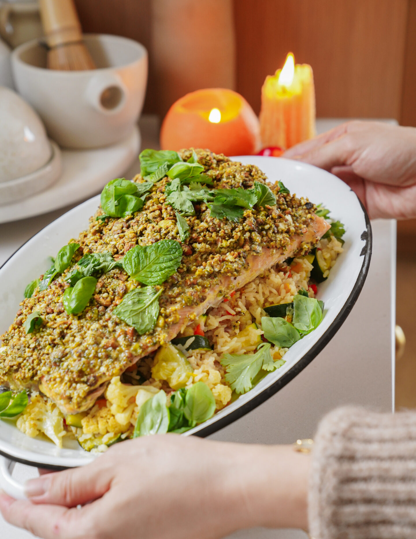 A person holds a platter with a pistachio-crusted salmon fillet on top of saffron rice and cauliflower. Fresh basil leaves garnish the dish. In the background, candles and a kitchen counter with jars are visible.