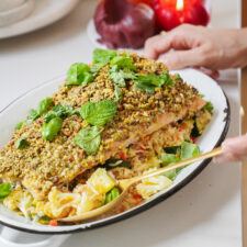 A hand places a spoon beside a dish of pistachio-crusted salmon on a bed of vegetable rice. Fresh basil garnishes the dish. In the background, a red onion and a red candle can be seen on the countertop.