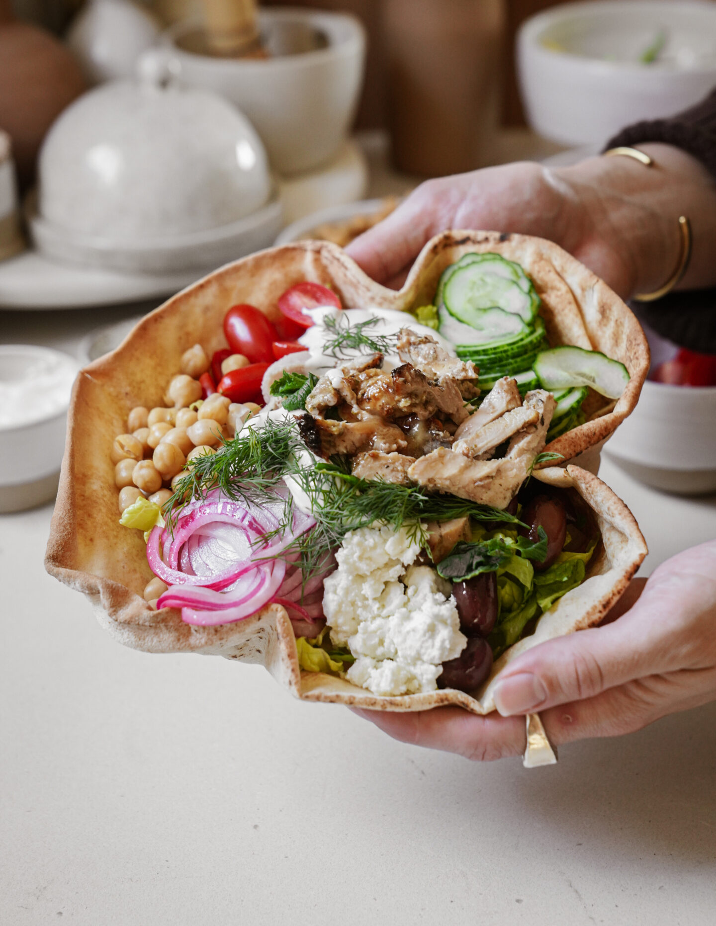 A persons hands holding a pita bowl filled with grilled chicken, sliced cucumbers, cherry tomatoes, chickpeas, pickled onions, feta cheese, lettuce, and garnished with dill. Bowls and mortar sets are visible in the blurred background.