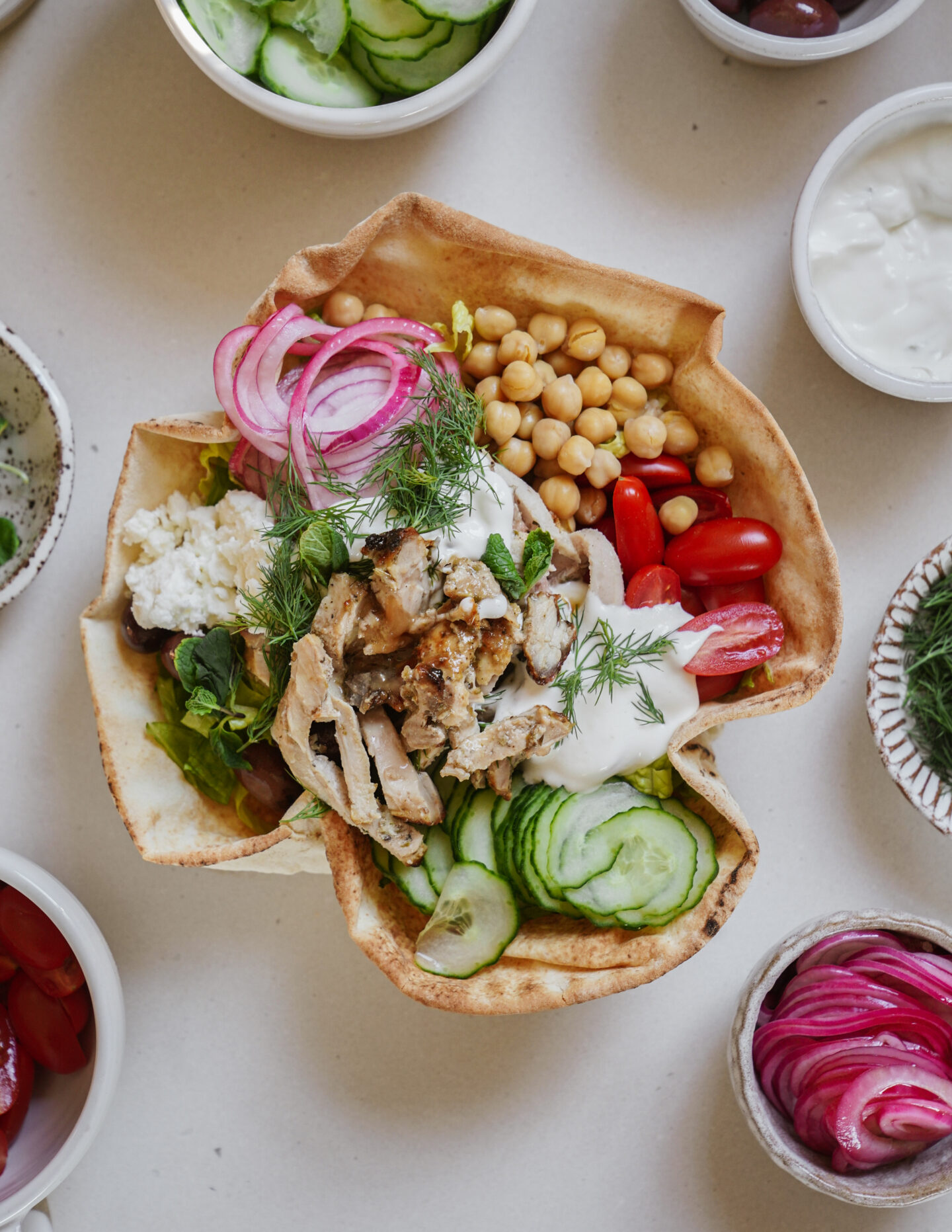 A bowl made from pita bread filled with grilled chicken, chickpeas, cucumber slices, cherry tomatoes, red onions, and dollops of yogurt sauce, garnished with dill. Surrounding bowls contain cucumbers, yogurt, and pickled onions.