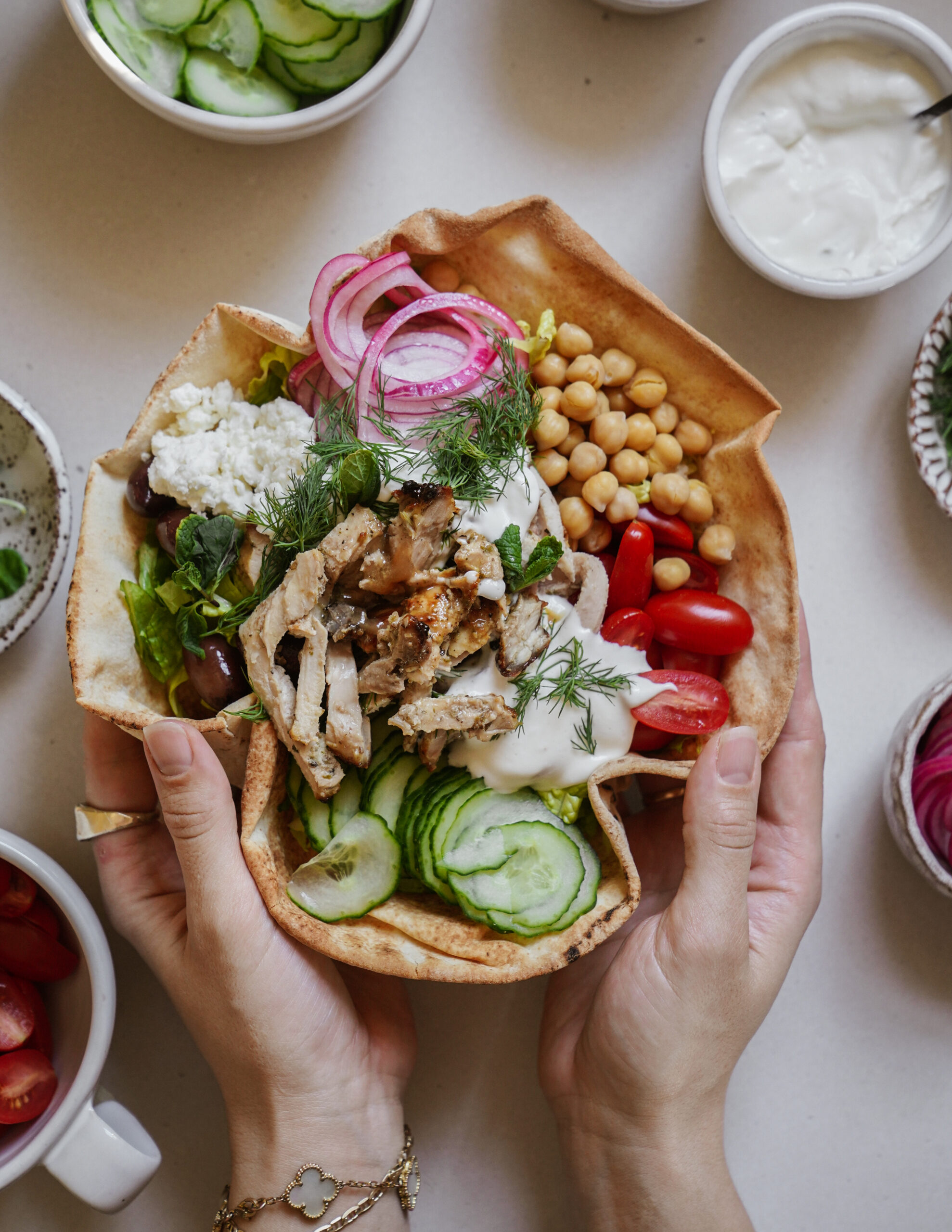 Hands holding a pita filled with sliced grilled chicken, cucumber, tomatoes, chickpeas, lettuce, feta cheese, olives, pickled onions, and yogurt sauce. Surrounded by bowls of sliced vegetables and yogurt on a light surface.