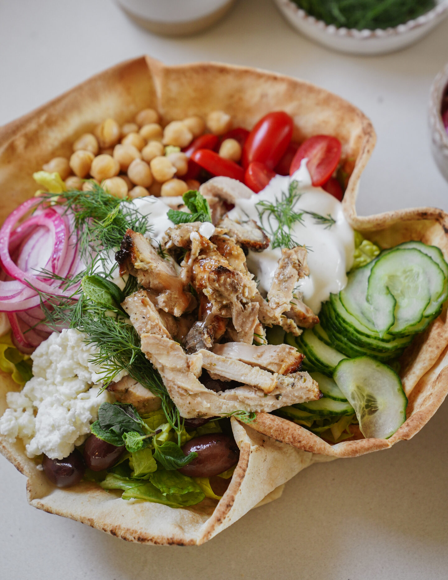 Pita bowl filled with grilled chicken, sliced cucumbers, halved cherry tomatoes, chickpeas, pickled red onions, crumbled feta, yogurt sauce, fresh dill, and olives, all on a bed of lettuce.