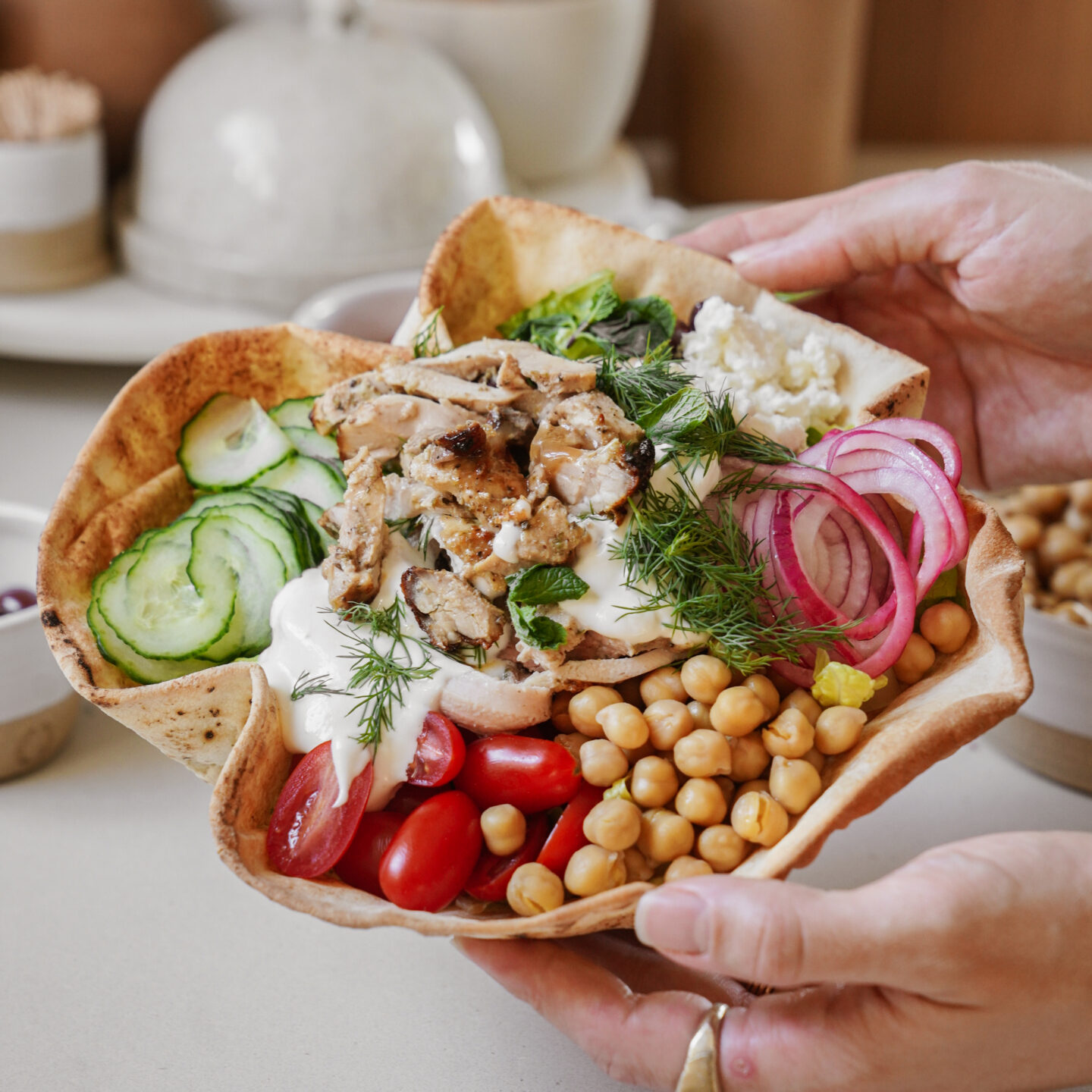 A person holding a bowl made from pita bread filled with sliced grilled chicken, chickpeas, cucumber slices, cherry tomatoes, pickled red onions, and feta cheese, garnished with fresh dill.