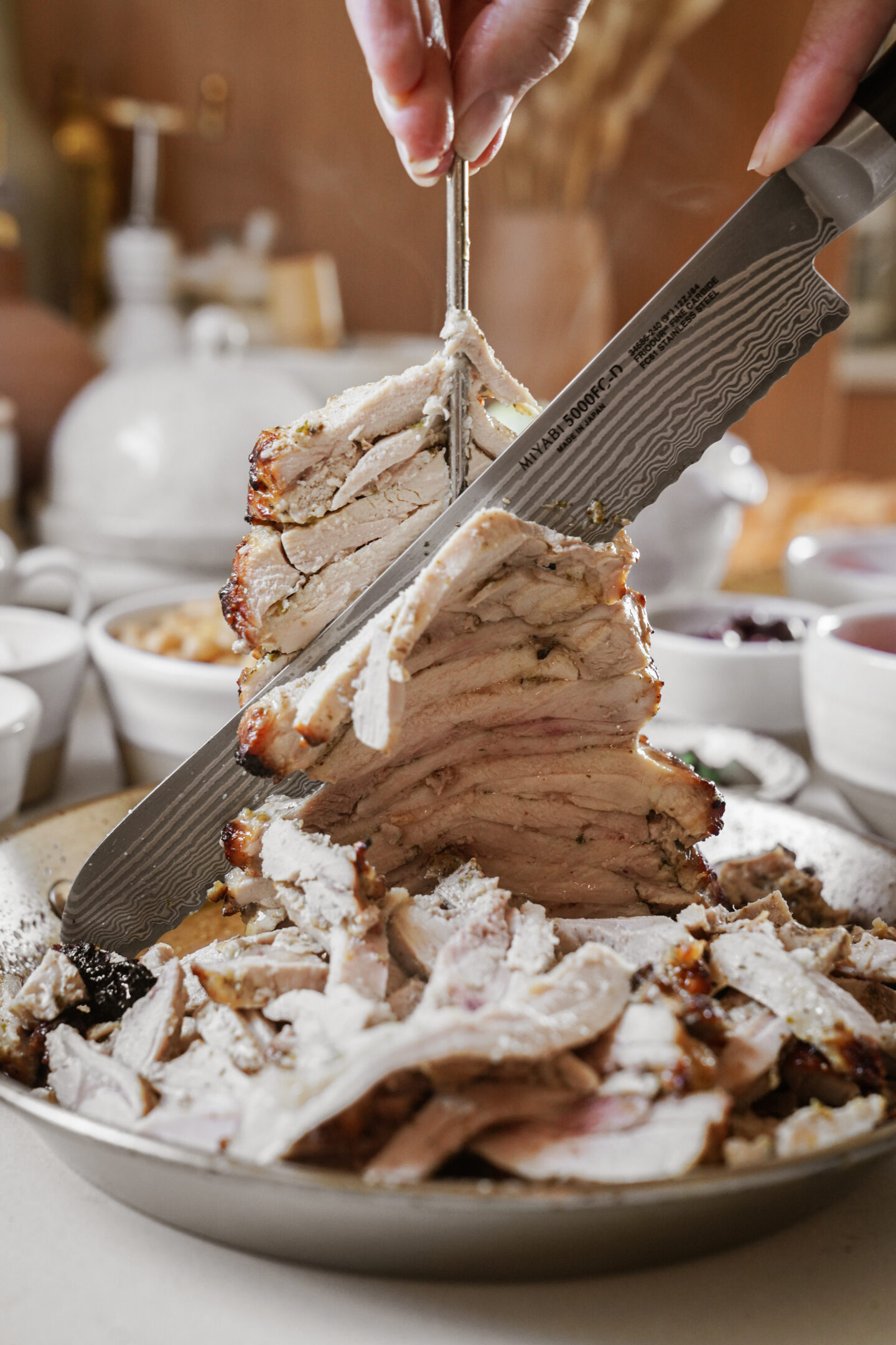 A hand uses a knife and fork to carve juicy slices from a stack of roasted meat on a plate. The sliced meat appears tender and is surrounded by various small bowls in the background.